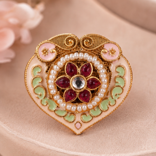 An ornate heart-shaped brooch with gold-plated detailing, pink enamel, light green enamel, white faux pearls, and a central red floral motif with a clear center stone, displayed on a beige surface.
