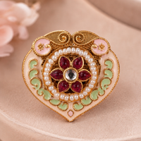 An ornate heart-shaped brooch with gold-plated detailing, pink enamel, light green enamel, white faux pearls, and a central red floral motif with a clear center stone, displayed on a beige surface.