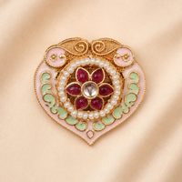An ornate heart-shaped brooch featuring gold-plated metalwork, pink enamel, light green enamel, white faux pearls, and a central cluster of red gemstones, displayed on a soft beige satin background.