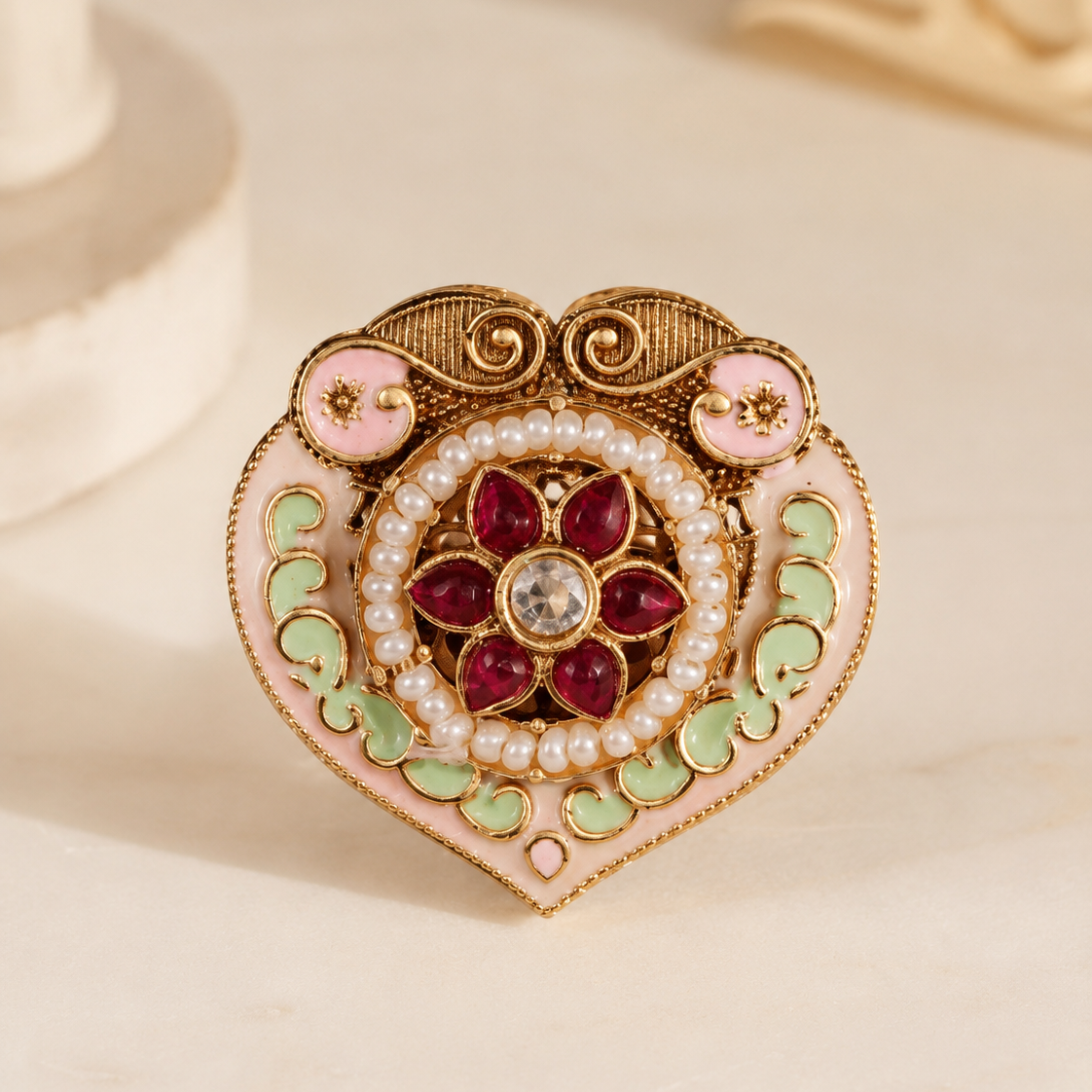 An ornate heart-shaped brooch with gold-tone filigree, pink enamel, green enamel, white faux pearls, and red gemstone petals, photographed on a light beige surface.