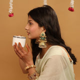 A woman in profile wearing a light cream-colored traditional outfit and ornate gold-toned jewelry, holding a small white cup against a warm beige background with hanging floral decorations.