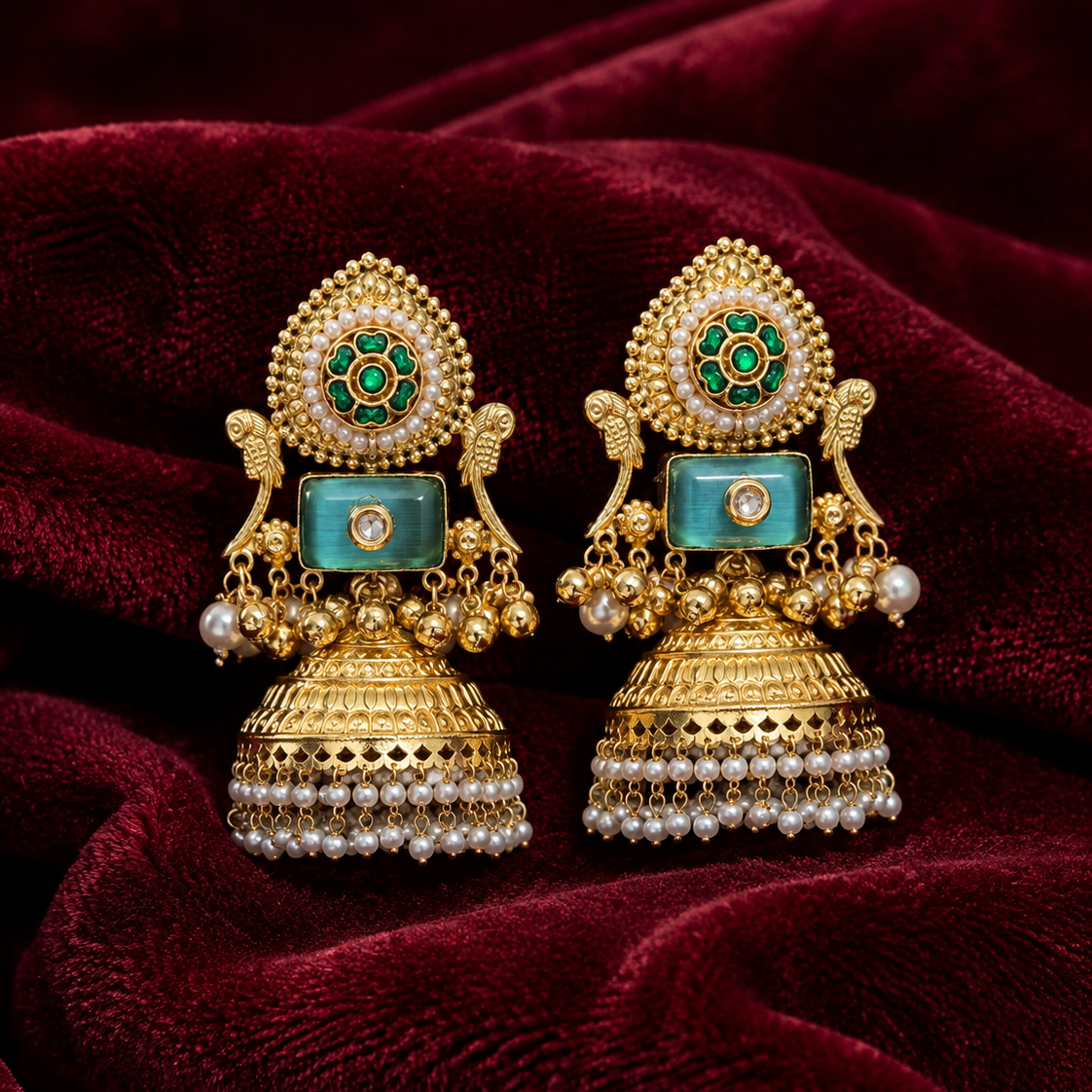 A pair of ornate gold-toned traditional earrings with turquoise blue stone inlays and green enamel details, displayed on a deep burgundy velvet fabric background.
