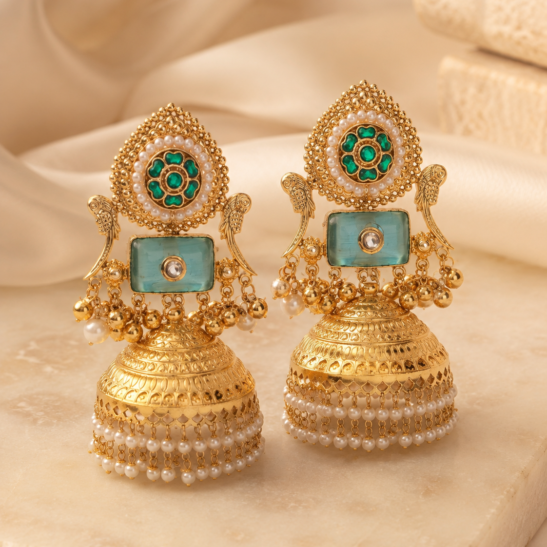 A pair of traditional Indian gold-plated jhumka earrings with green and white stone accents, displayed on a beige marble surface with a soft beige fabric background.