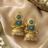 A pair of ornate gold-toned Indian jhumka earrings with turquoise blue rectangular stones, green and white enamel details, and white pearl bead fringe, displayed on draped champagne-colored satin fabric.