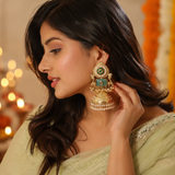 A woman in a light sage green saree wearing a large, ornate gold-toned traditional Indian jhumka earring with green and white stones, shown in a warm, softly lit indoor setting.