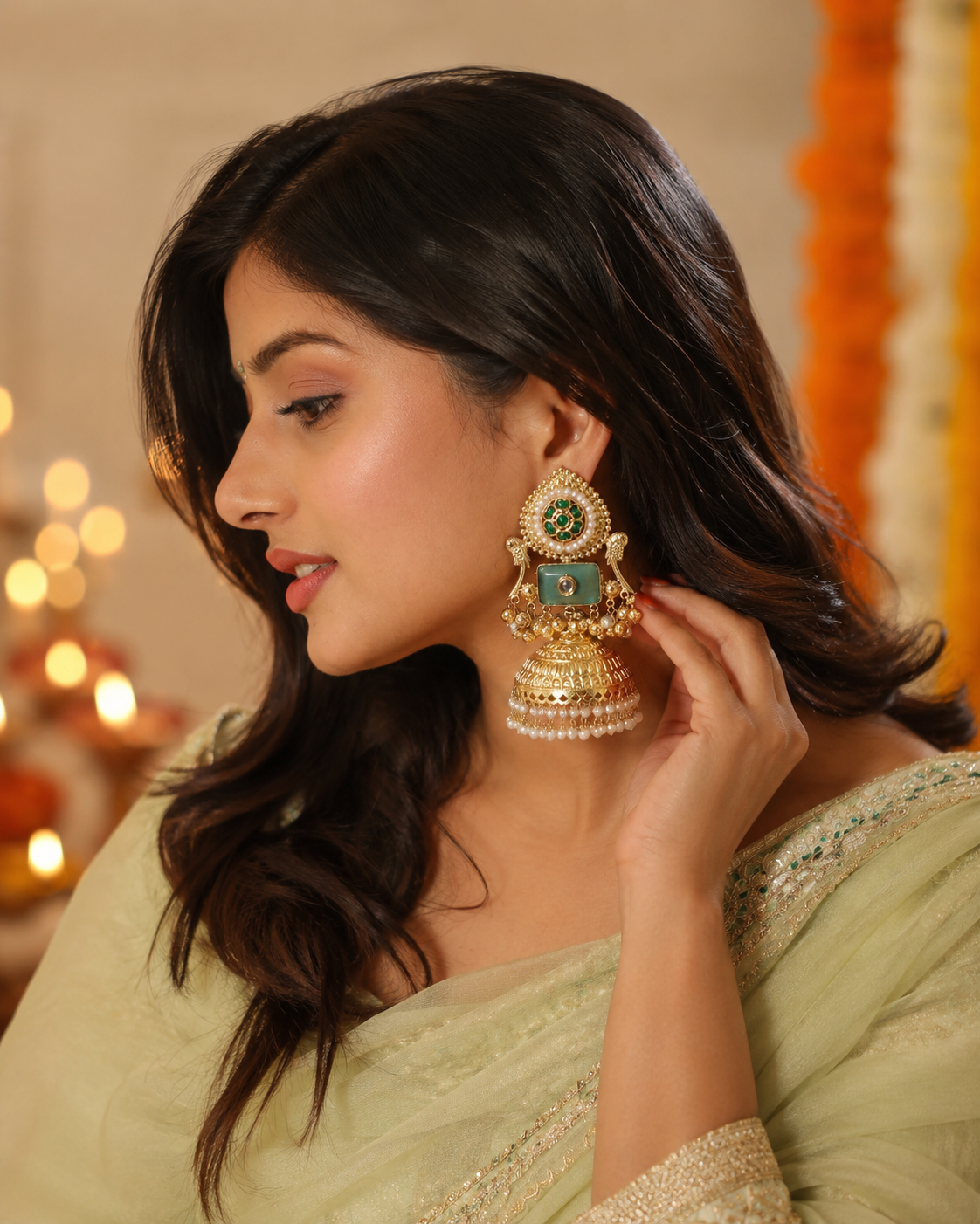 A woman in a light sage green saree wearing a large, ornate gold-toned traditional Indian jhumka earring with green and white stones, shown in a warm, softly lit indoor setting.