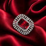 A square brooch featuring a central rectangular red gemstone surrounded by a double halo of small clear stones in a silver-toned setting, displayed on draped red satin fabric.