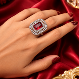 A close-up of a hand wearing a large rectangular red gemstone ring with a halo of small clear stones, set against a rich red satin fabric background.